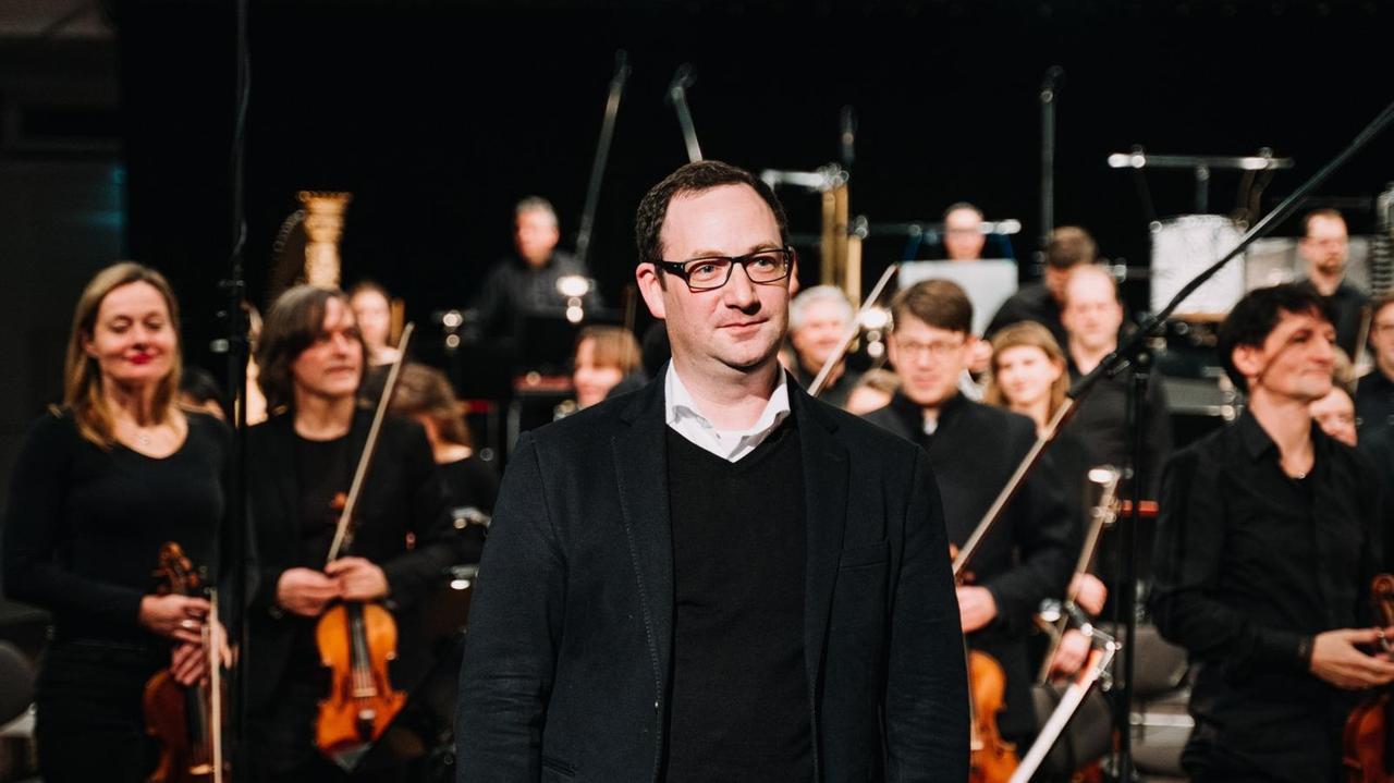 Der Komponist steht vor Orchestermusikerinnen und nimmt den Beifall entgegen. Der Komponist steht vor Orchestermusikerinnen und nimmt den Beifall entgegen.