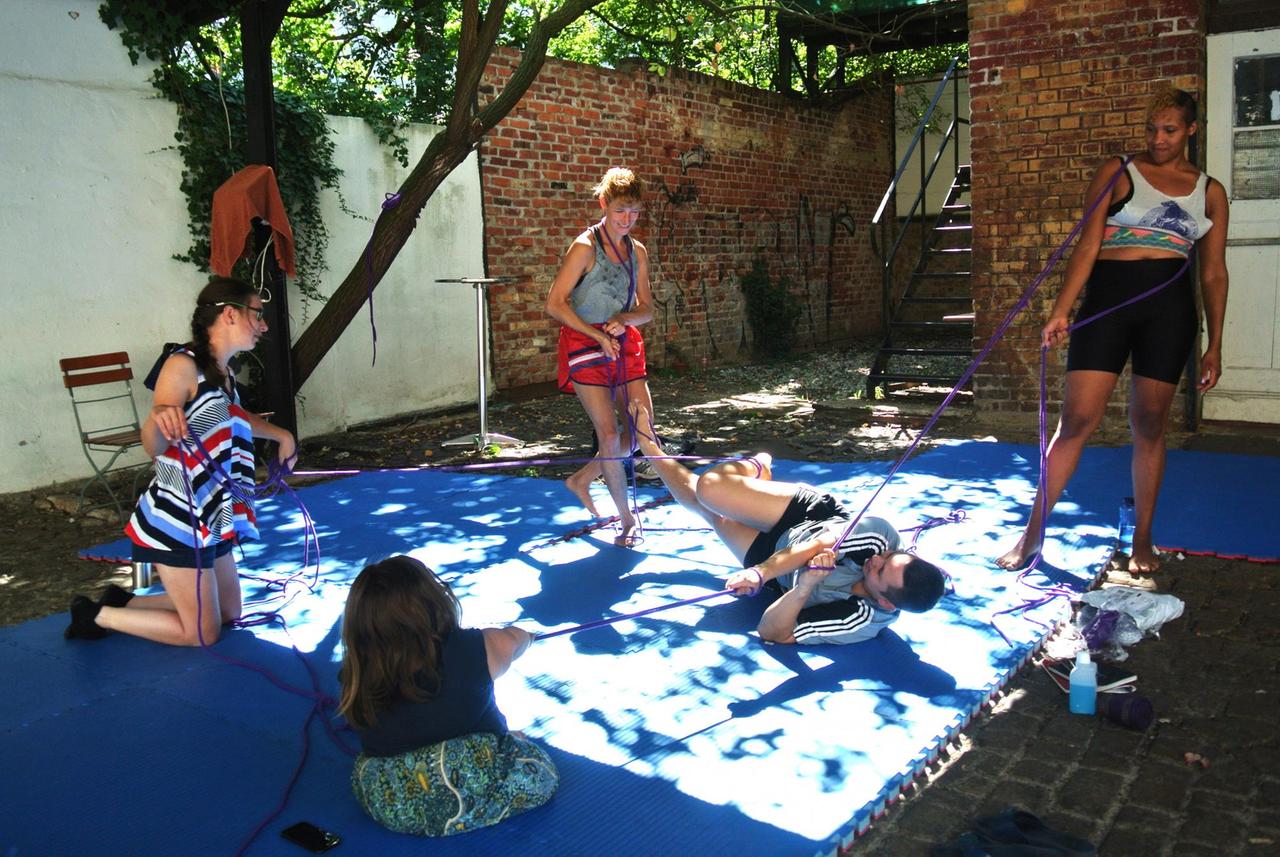 Das Performancekollektiv Polymora Inc bei der Probe unter freiem Himmel. 5 Personen halten sich an einem Seil fest und sind somit alle miteinander verbunden. Die Person in der Mitte liegt auf dem Boden, als wäre sie die Beute der anderen. Das Performancekollektiv Polymora Inc bei der Probe unter freiem Himmel. 5 Personen halten sich an einem Seil fest und sind somit alle miteinander verbunden. Die Person in der Mitte liegt auf dem Boden, als wäre sie die Beute der anderen.