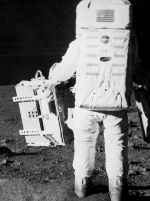 Der Astronaut Edwin E. Aldrin Jr. läuft über die Mondoberfläche und trägt zwei Taschen mit wissenschaftlichem Equipment bei sich.