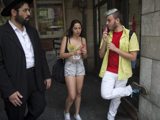 Ein orthodoxer jüdischer Mann (li.) geht in Jerusalem an zwei modern gekleideten israelischen Jugendlichen vorbei, die an einer Demonstration gegen sexuelle Belästigung teilnehmen. Die Demonstration steht unter dem Motto "Sagt nicht uns, was wir tragen sollen - sondern sagt ihnen, dass sie uns nicht vergewaltigen sollen".