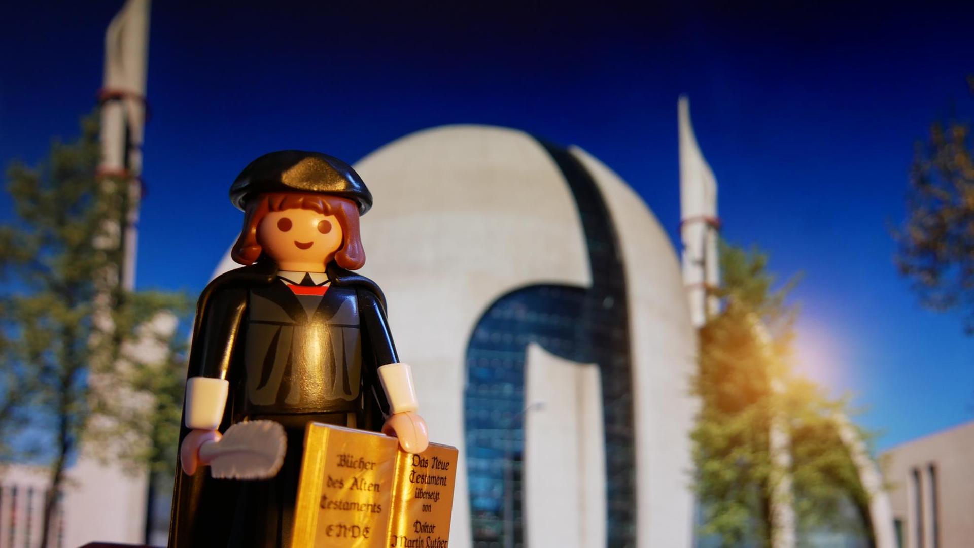Was würde Luther zur Kölner Moschee sagen? Ein Playmobilmännchen, das als Martin Luther zurechtgemacht ist, steht vor der Kölner Moschee und hält eine Feder und die Lutherbibel in den Händen.