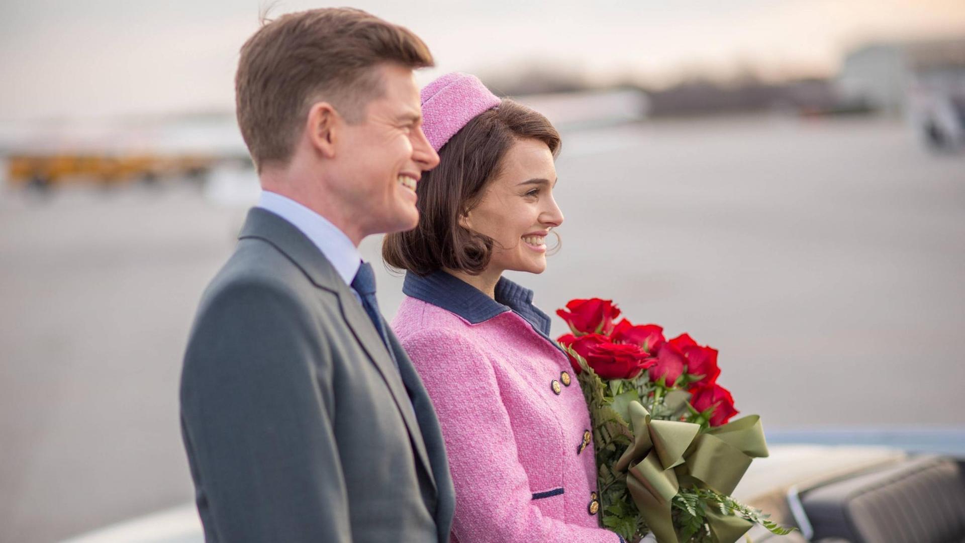John F. Kennedy (Caspar Phillipson) steht neben seiner Frau Jackie (Natalie Portman), die einen Strauß rote Rosen in der Hand hält. Beide sind im Profil zu sehen und lächeln