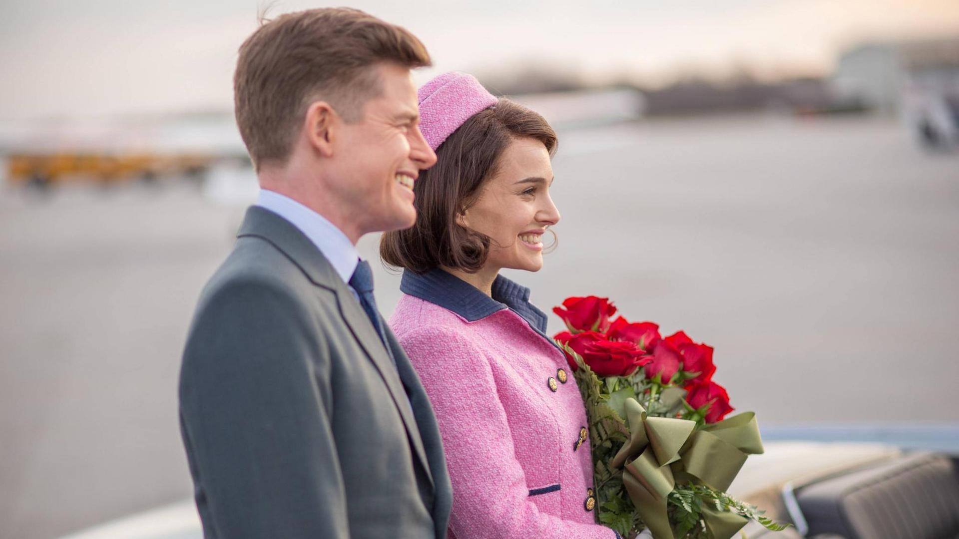 John F. Kennedy (Caspar Phillipson) steht neben seiner Frau Jackie (Natalie Portman), die einen Strauß rote Rosen in der Hand hält. Beide sind im Profil zu sehen und lächeln John F. Kennedy (Caspar Phillipson) steht neben seiner Frau Jackie (Natalie Portman), die einen Strauß rote Rosen in der Hand hält. Beide sind im Profil zu sehen und lächeln