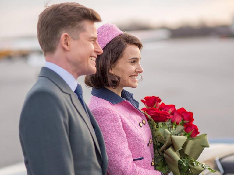 John F. Kennedy (Caspar Phillipson) steht neben seiner Frau Jackie (Natalie Portman), die einen Strauß rote Rosen in der Hand hält. Beide sind im Profil zu sehen und lächeln