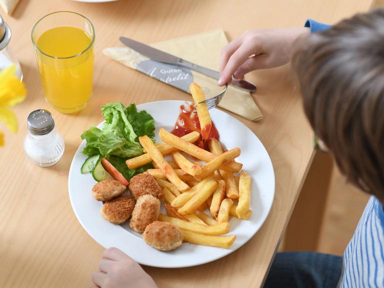 Vor einem Teller sitzt ein Kind mit Pommes und Chicken-Nuggets.