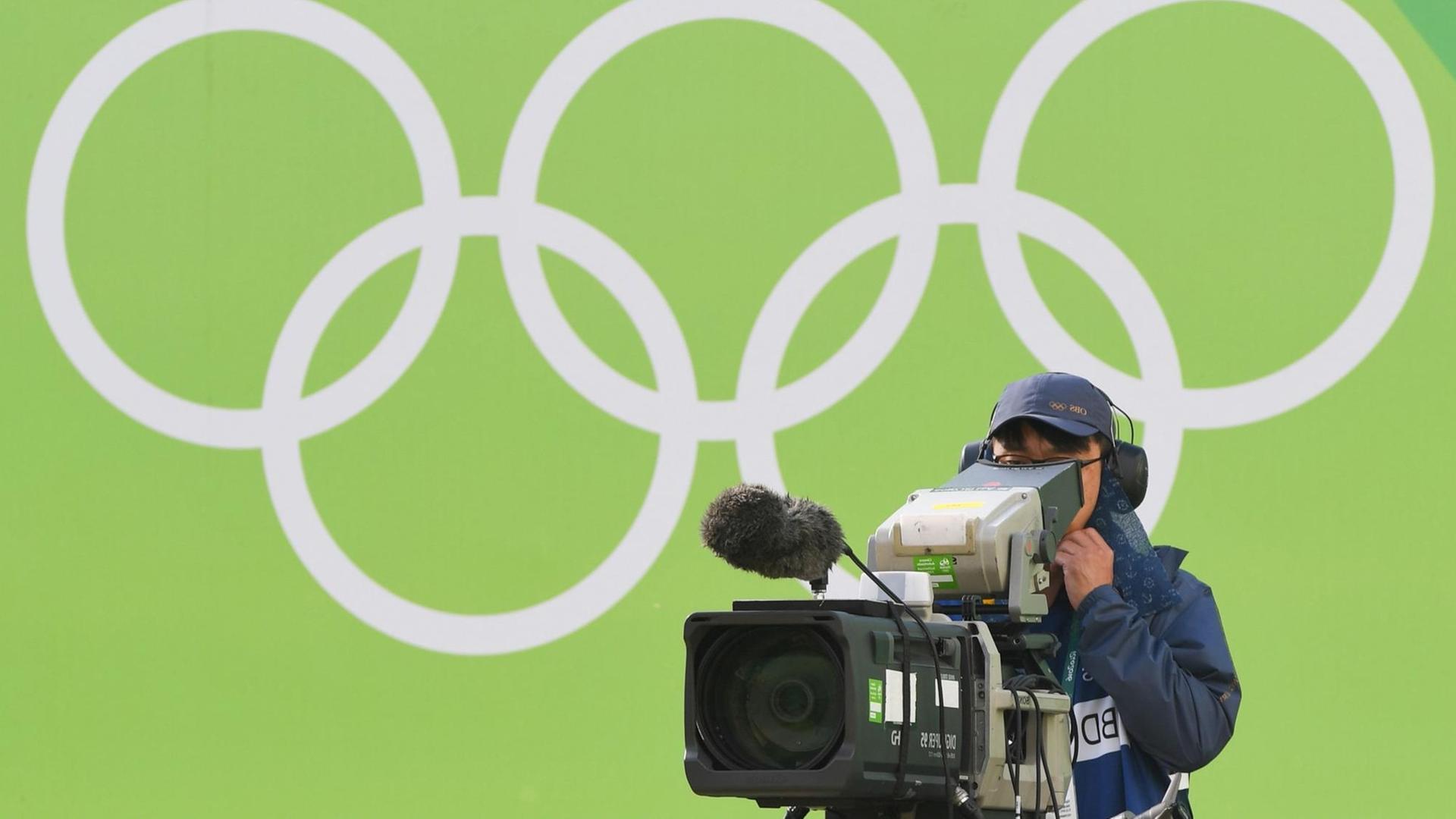 Ein TV-Kameramann filmt bei den Olympischen Spielen 2016 in Rio, hinter ihm sind die Olympischen Ringe zu sehen. Ein TV-Kameramann filmt bei den Olympischen Spielen 2016 in Rio, hinter ihm sind die Olympischen Ringe zu sehen.