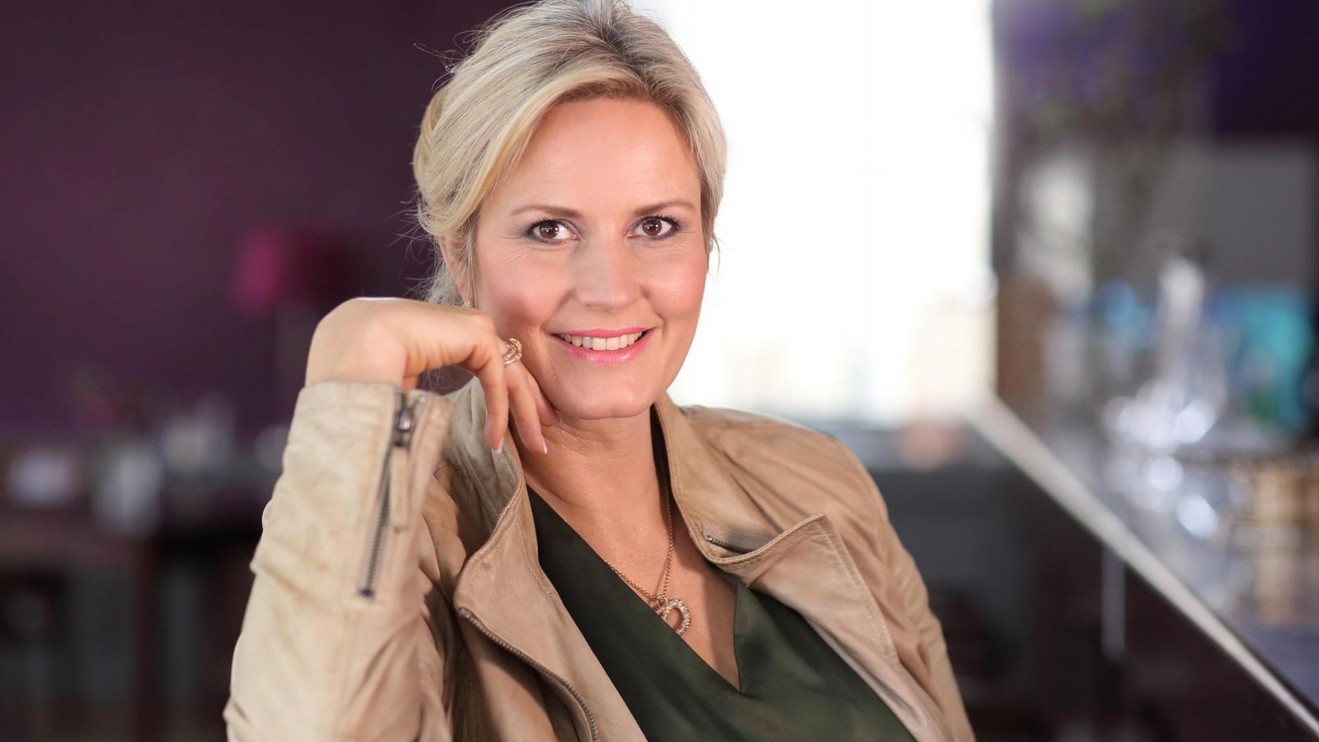 Eine Frau mit langen blonden Haaren sitzt zurückgelehnt und schaut auf einer Halbporträtaufnahme frontal in die Kamera. Sie lächelt und hält ihre rechte Hand an das Gesicht. Eine Frau mit langen blonden Haaren sitzt zurückgelehnt und schaut auf einer Halbporträtaufnahme frontal in die Kamera. Sie lächelt und hält ihre rechte Hand an das Gesicht.