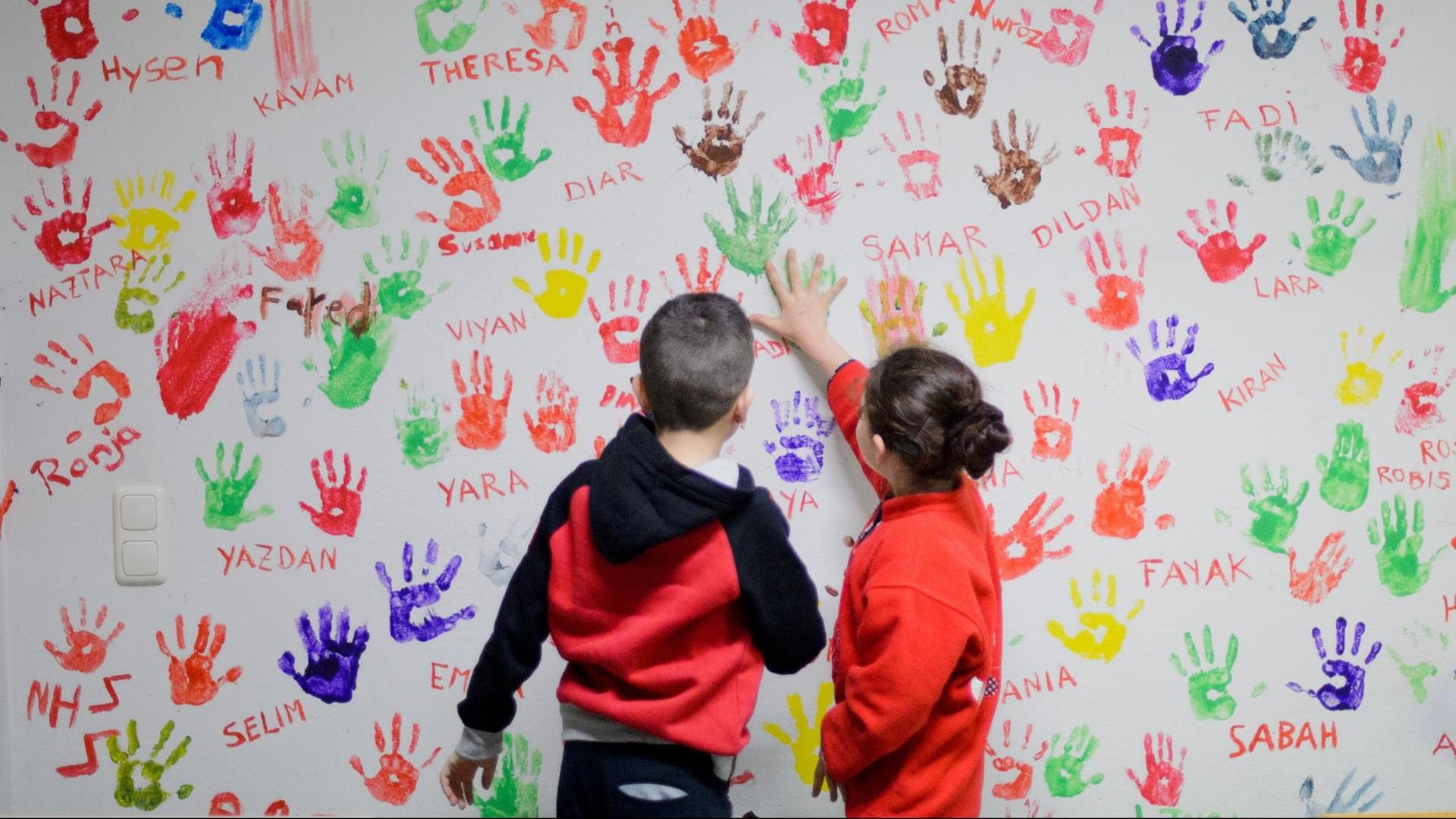 Zwei Flüchtlingskinder in einer Erstaufnahmeeinrichtung für Flüchtlinge in Hameln vor einer Wand, auf der sich junge Flüchtlinge aus der ganzen Welt mit bunten Handabdrücken verewigt haben. Zwei Flüchtlingskinder in einer Erstaufnahmeeinrichtung für Flüchtlinge in Hameln vor einer Wand, auf der sich junge Flüchtlinge aus der ganzen Welt mit bunten Handabdrücken verewigt haben.