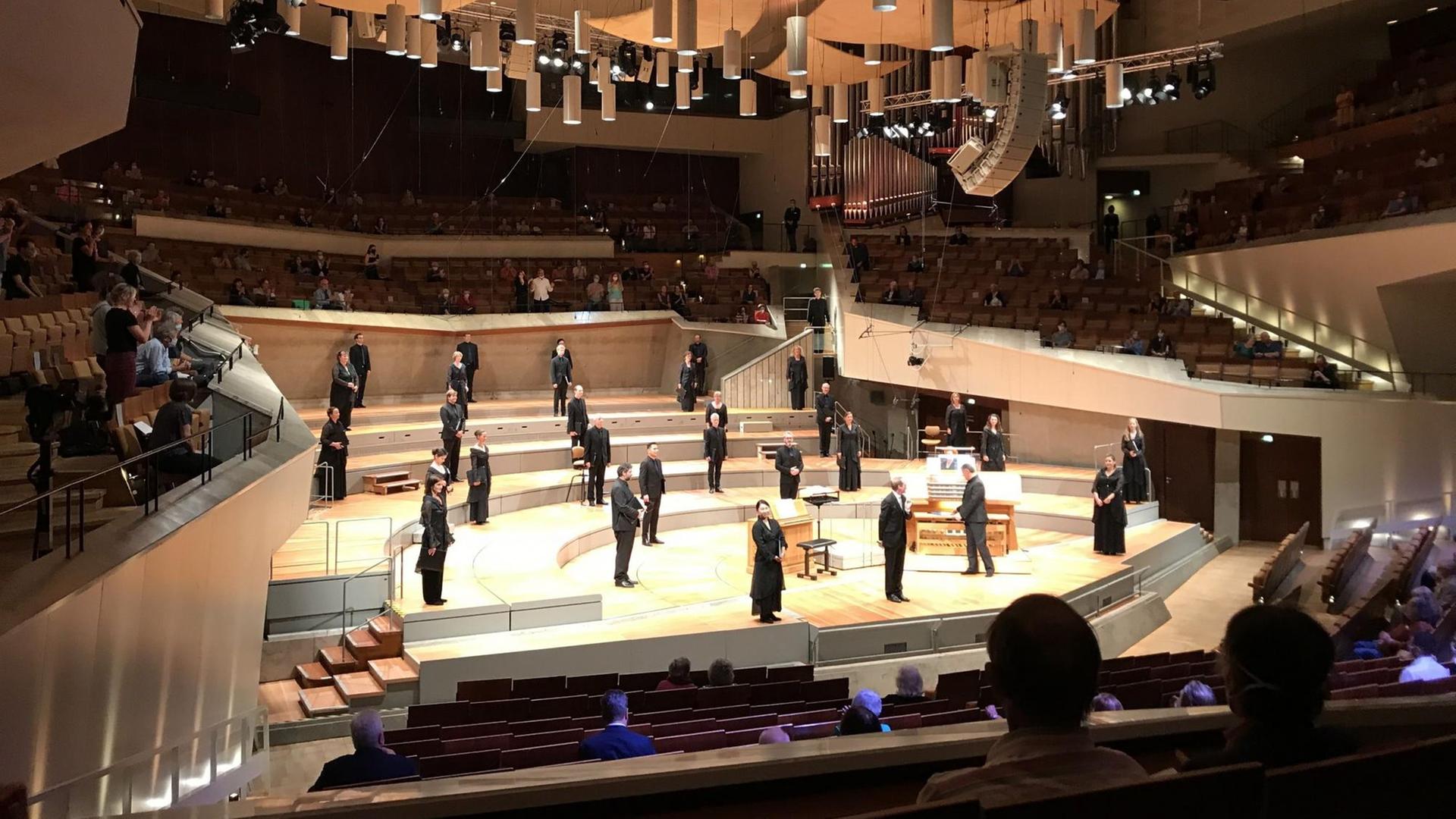 Es ist ein Konzertsaal zu sehen mit einem auf Abstand stehendem Chor auf der Bühne und rar besetzten Publikumssitzen Es ist ein Konzertsaal zu sehen mit einem auf Abstand stehendem Chor auf der Bühne und rar besetzten Publikumssitzen