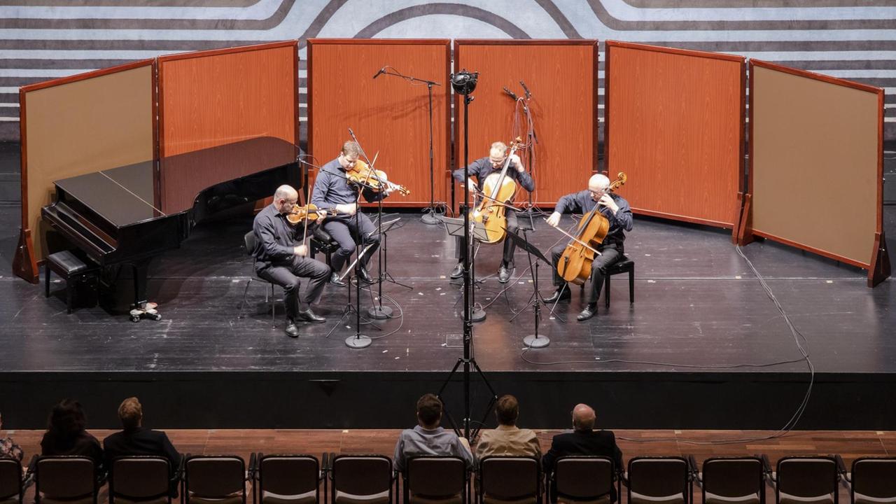 Auf einer Bühne, die mit orangefarbenen Wänden im Halbkreis betont wird, spielen vier Musiker ihre Streichinstrumente. Auf einer Bühne, die mit orangefarbenen Wänden im Halbkreis betont wird, spielen vier Musiker ihre Streichinstrumente.