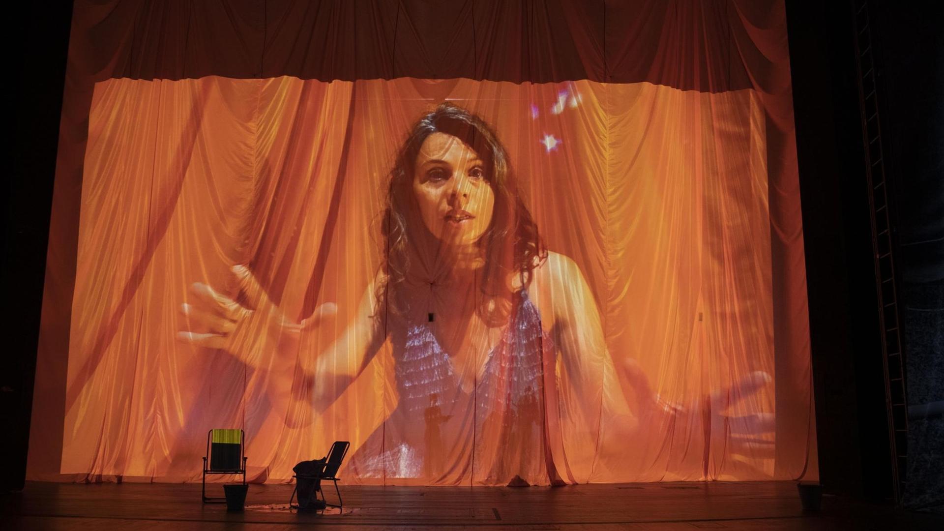 Auf einen Vorhang auf der Bühne im Berliner Theater Volksbühne wird ein Bild der Schauspielerin Margarita Breitkreiz projiziert. Die Frau mit dunklen, langen Haaren gestikuliert, die Falten des Vorhangs verfremden das Bild. Vor dem Vorhang stehen zwei Stühle. Auf einen Vorhang auf der Bühne im Berliner Theater Volksbühne wird ein Bild der Schauspielerin Margarita Breitkreiz projiziert. Die Frau mit dunklen, langen Haaren gestikuliert, die Falten des Vorhangs verfremden das Bild. Vor dem Vorhang stehen zwei Stühle.