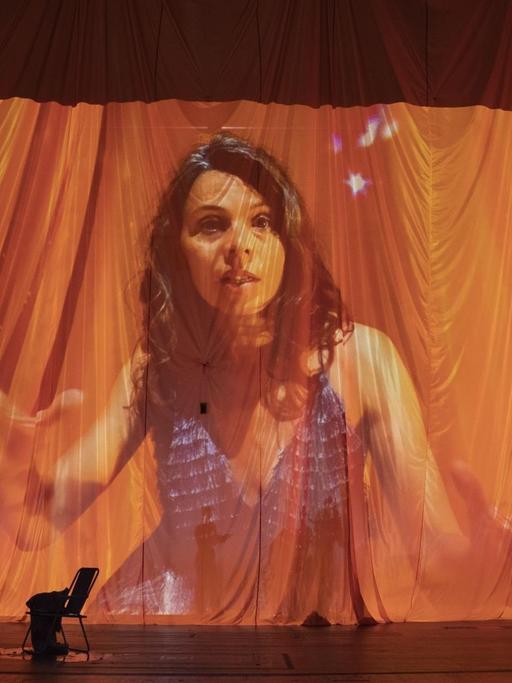 Auf einen Vorhang auf der Bühne im Berliner Theater Volksbühne wird ein Bild der Schauspielerin Margarita Breitkreiz projiziert. Die Frau mit dunklen, langen Haaren gestikuliert, die Falten des Vorhangs verfremden das Bild. Vor dem Vorhang stehen zwei Stühle.