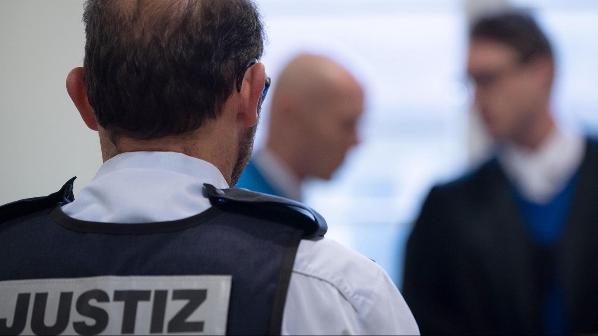 Das Bild zeigt den Kopf des Angeklagten unscharf im Hintergrund neben seinem Verteidiger. Im Vordergrund scharf der Rücken eines Justizbeamten mit der Rückenaufschrift "Justiz". Das Bild zeigt den Kopf des Angeklagten unscharf im Hintergrund neben seinem Verteidiger. Im Vordergrund scharf der Rücken eines Justizbeamten mit der Rückenaufschrift "Justiz".