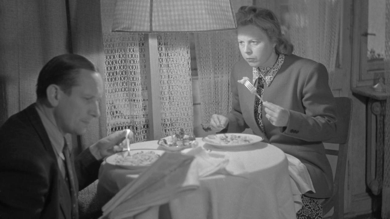 Mann und Frau sitzen gemeinsam beim Essen an einem kleinen Tisch, der Mann liest und die Frau sieht ihn leicht verärgert an. Mann und Frau sitzen gemeinsam beim Essen an einem kleinen Tisch, der Mann liest und die Frau sieht ihn leicht verärgert an.