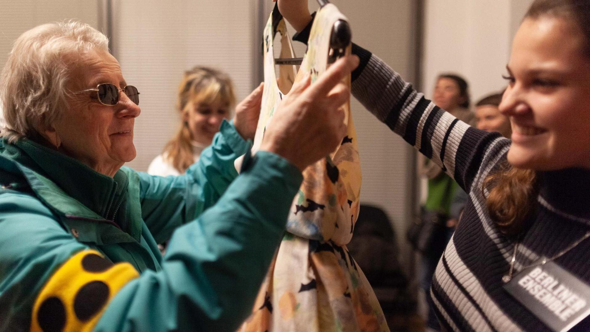 Eine Frau mit dem Blindensymbol tastet an einem Kleidungsstück, das von einer Mitarbeiterin des Berliner Ensembles hochgehalten wird. Eine Frau mit dem Blindensymbol tastet an einem Kleidungsstück, das von einer Mitarbeiterin des Berliner Ensembles hochgehalten wird.