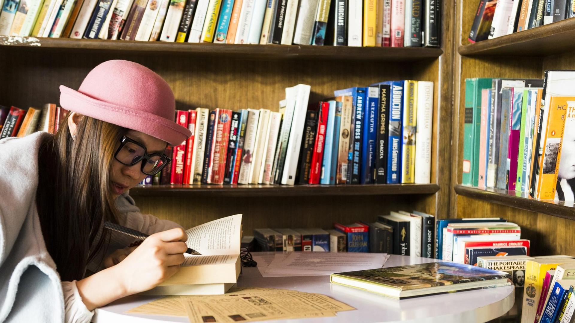 Ein junge Frau, mit einem aufgeschlagenem Buch in den Händen, sitzt am Tisch vor einem vollen Bücherregal. Ein junge Frau, mit einem aufgeschlagenem Buch in den Händen, sitzt am Tisch vor einem vollen Bücherregal.