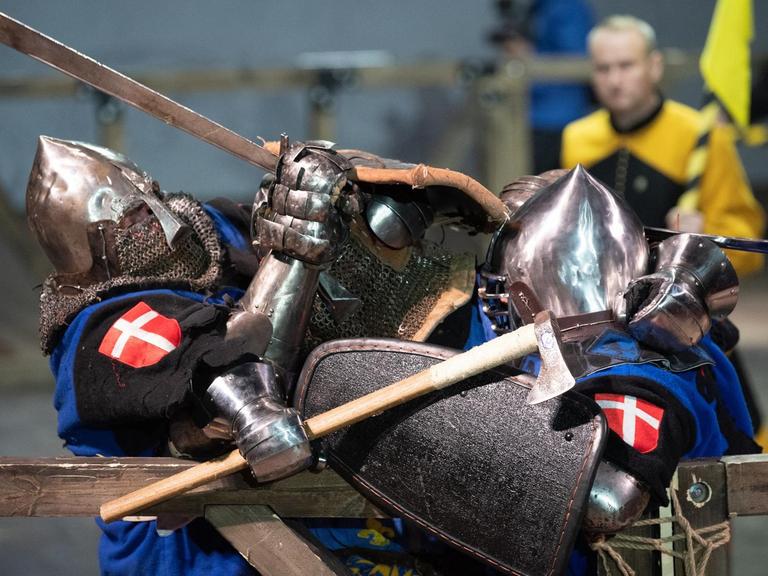 Zwei Kämpfer in mittelalterlicher Rüstung, mit Schild, Helm und Schwert.