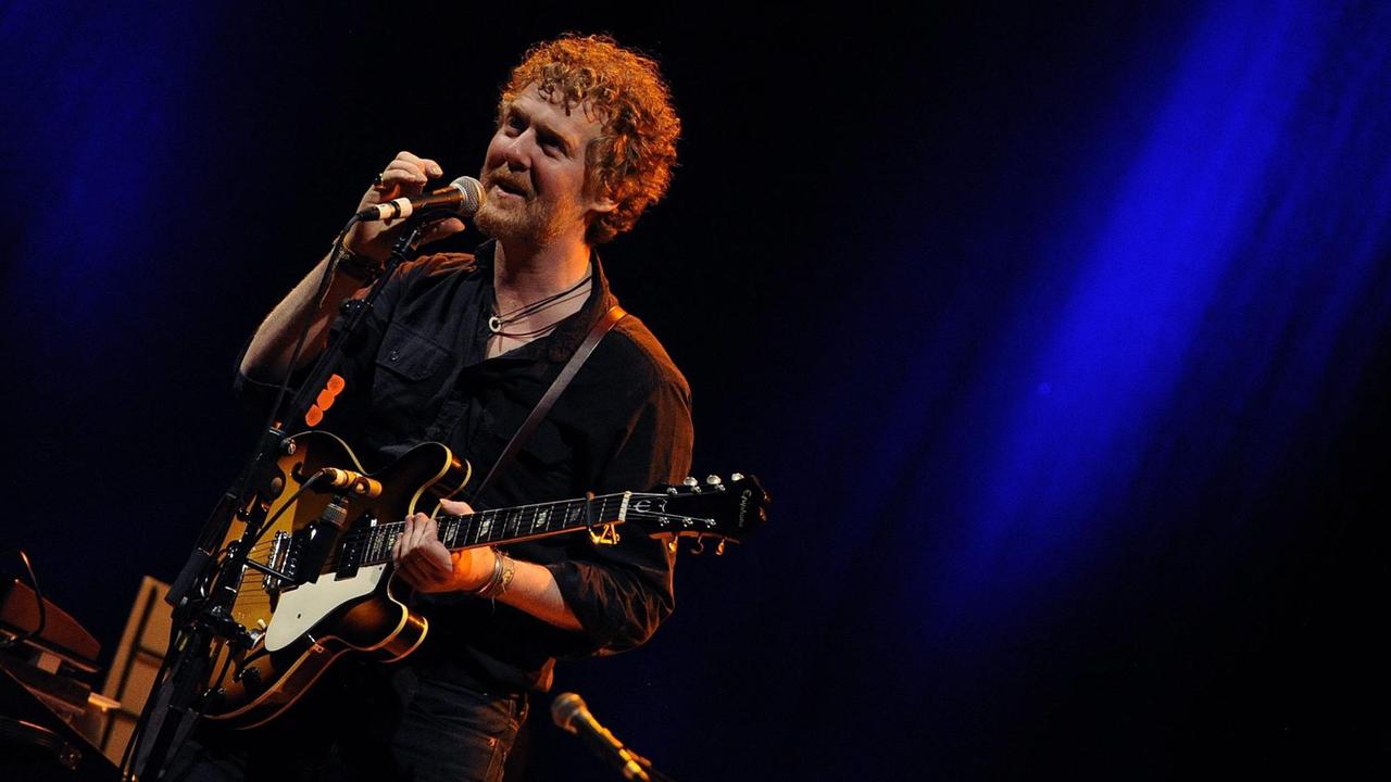 Der Musiker Glen Hansard auf einer dunklen Bühne stehend, mit einer Gitarre, in ein Mikrofon singend Der Musiker Glen Hansard auf einer dunklen Bühne stehend, mit einer Gitarre, in ein Mikrofon singend