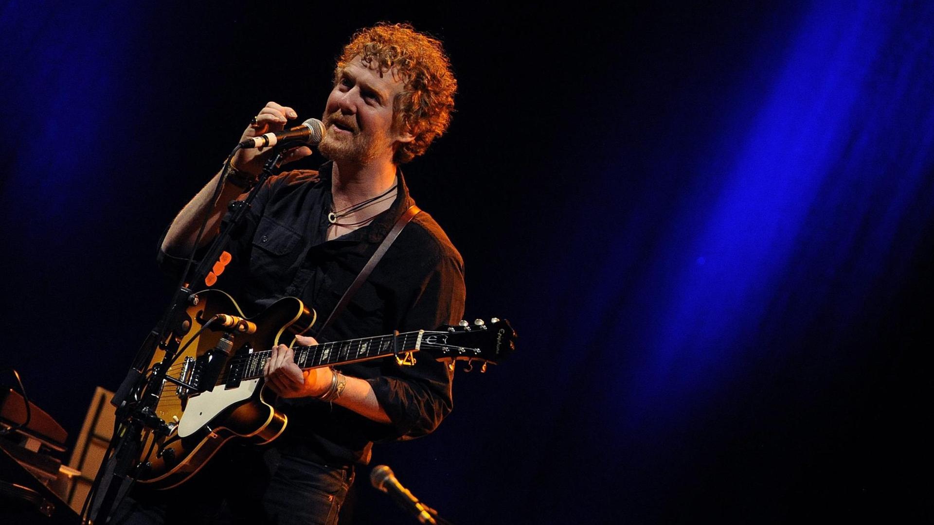 Der Musiker Glen Hansard auf einer dunklen Bühne stehend, mit einer Gitarre, in ein Mikrofon singend Der Musiker Glen Hansard auf einer dunklen Bühne stehend, mit einer Gitarre, in ein Mikrofon singend
