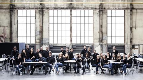 Männer und Frauen des Chores sitzen in blauen Jeans und schwarzen Oberteilen auf Stühlen in einer Werkhalle mit großen Fenstern.
