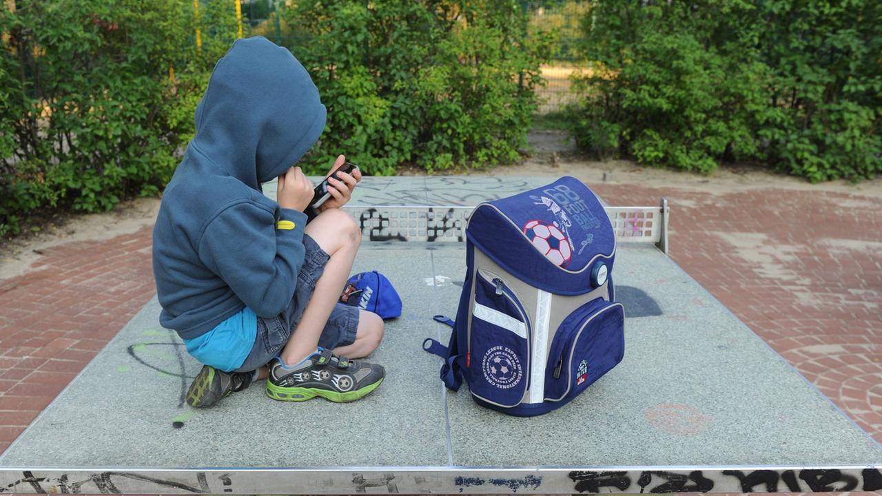 Ein Junge sitzt mit seinem Schulranzen auf einer Tischtennisplatte auf einem Spielplatz in Berlin und spielt auf einem Smartphone Ein Junge sitzt mit seinem Schulranzen auf einer Tischtennisplatte auf einem Spielplatz in Berlin und spielt auf einem Smartphone