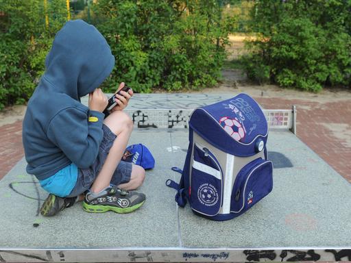 Ein Junge sitzt mit seinem Schulranzen auf einer Tischtennisplatte auf einem Spielplatz in Berlin und spielt auf einem Smartphone.