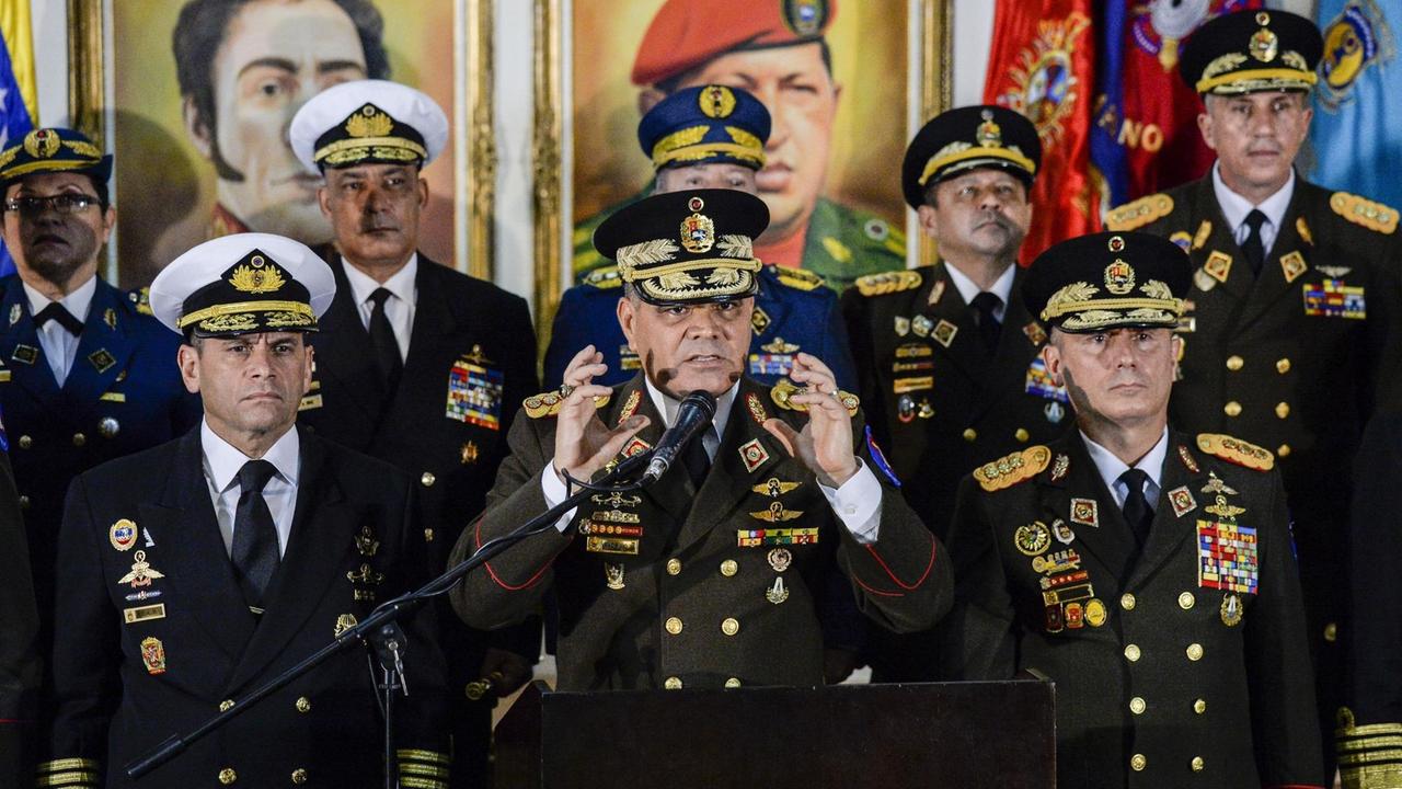 Das Bild zeigt Venezuelas Verteidigungsminister Vladimir Padrino gestikulierend während einer Erklärung hinter einem Mikrofon. Er trägt eine Uniform und ist umringt vonanderen Militärs. 