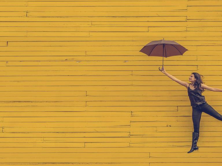 Vor einer gelb angemalten Holzwand springt eine Frau mit Regenschirm glücklich in die Luft.