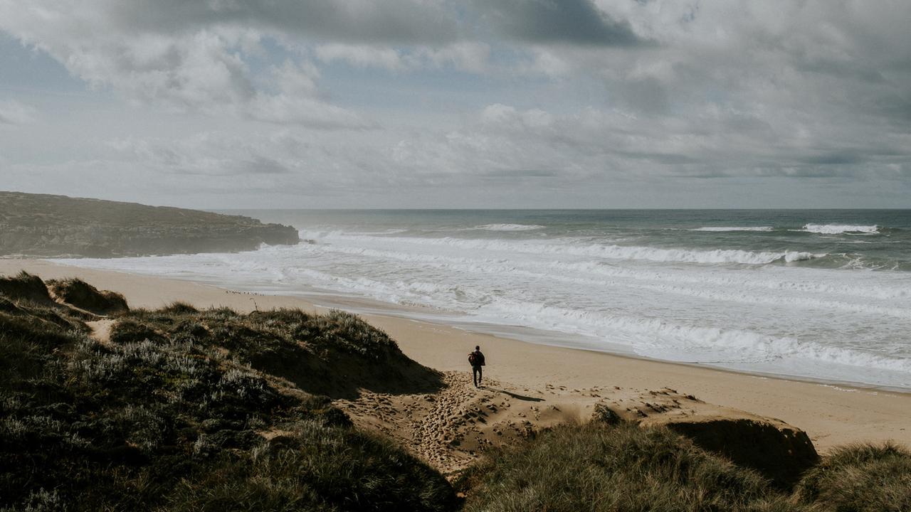 Ein Strand ist zu sehen, mit Felsen, Sand und bewölktem Himmel. Ein Mann von hinten geht in Richtung Wasser. Ein Strand ist zu sehen, mit Felsen, Sand und bewölktem Himmel. Ein Mann von hinten geht in Richtung Wasser.