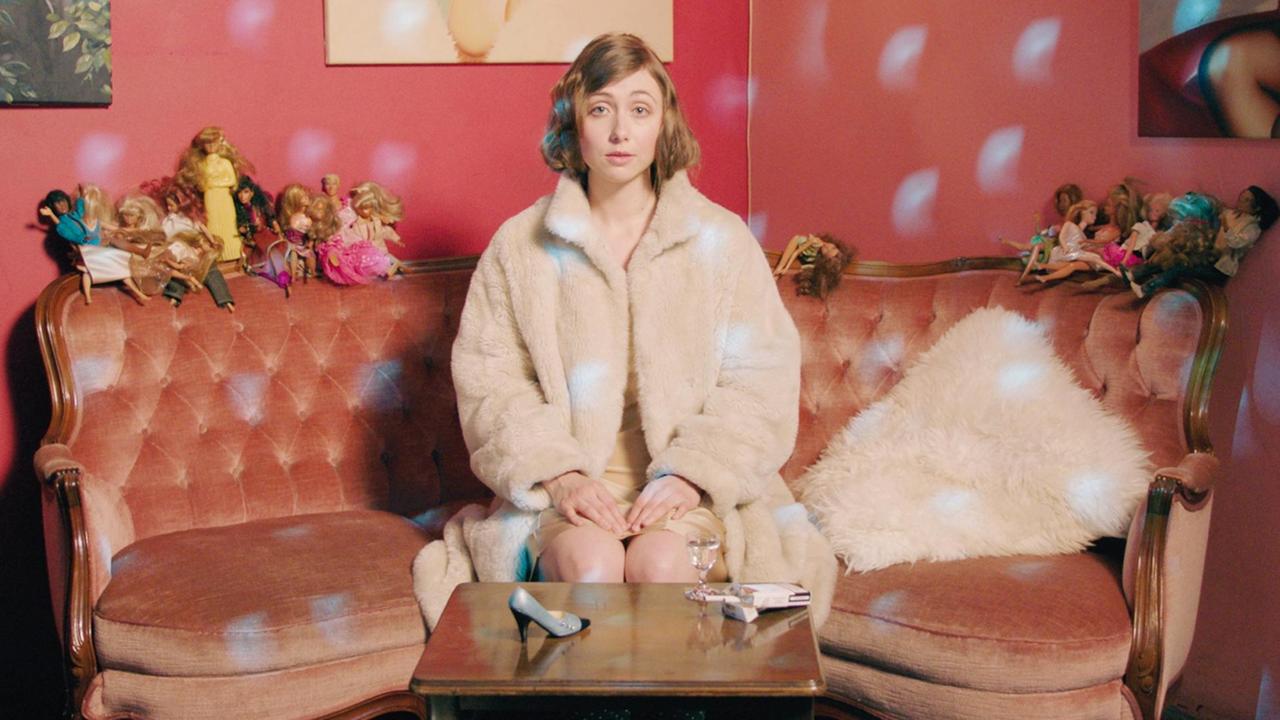 Marie Rathscheck sitzt auf einem altmodischen Sofa im Biedermeier-Stil und blickt den Betrachter an. Auf der Sofalehne befinden sich Mädchenpuppen im Barbie-Stil. Auf dem Tisch vor ihr steht ein einzelner Damenschuh, ein Glas mit Wasser und eine geöffnete Zigarettenschachtel. Marie Rathscheck sitzt auf einem altmodischen Sofa im Biedermeier-Stil und blickt den Betrachter an. Auf der Sofalehne befinden sich Mädchenpuppen im Barbie-Stil. Auf dem Tisch vor ihr steht ein einzelner Damenschuh, ein Glas mit Wasser und eine geöffnete Zigarettenschachtel.