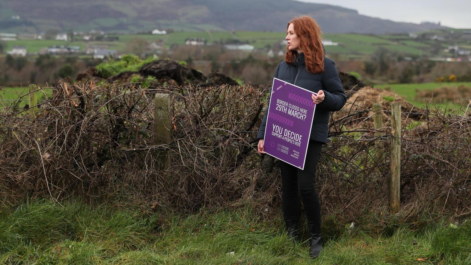 Eine Frau steht mitten in der Landschaft und protestiert mit einem Plakat an der irischen Grenze gegen den Brexit. Eine Frau steht mitten in der Landschaft und protestiert mit einem Plakat an der irischen Grenze gegen den Brexit.