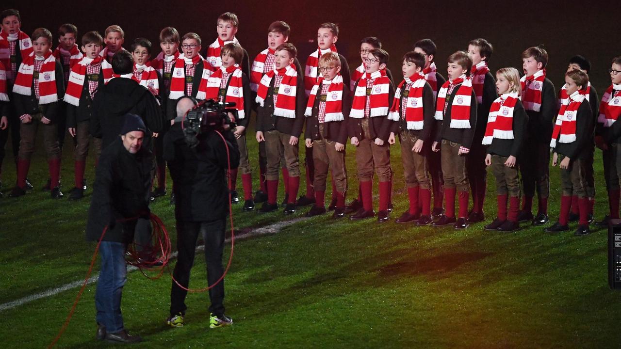 Die Jungen des Knabenchor stehen in zwei Reihen auf dem Fußballrasen und tragen taditionelle bayrische Kleidung und Schals in rot-weiß-gestreift, den Farben des FC Bayern München. Die Jungen des Knabenchor stehen in zwei Reihen auf dem Fußballrasen und tragen taditionelle bayrische Kleidung und Schals in rot-weiß-gestreift, den Farben des FC Bayern München.