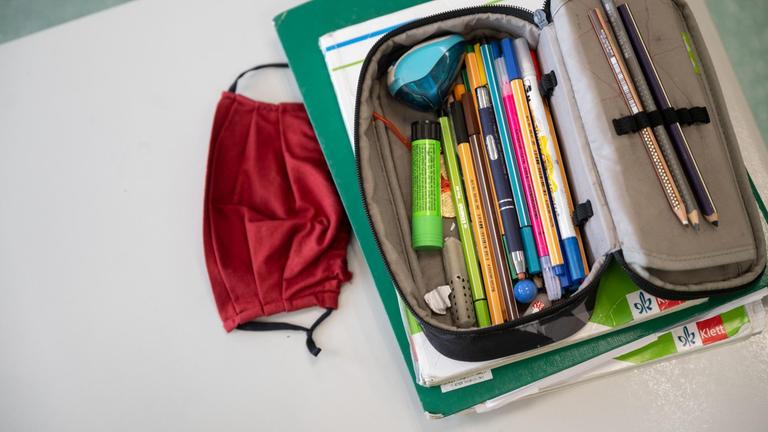 Eine Mund-Nasen-Bedeckung liegt während einer Unterrichtsstunde einer fünften Klasse des Friedrich-Schiller Gymnasiums neben einem Mäppchen und Schulbüchern auf einem Schultisch.