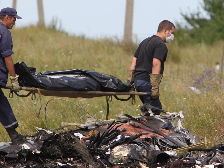 Hinter Trümmern tragen zwei ukrainische Arbeiter eine Bahre, auf der ein abgedecktes Todesopfer liegt.