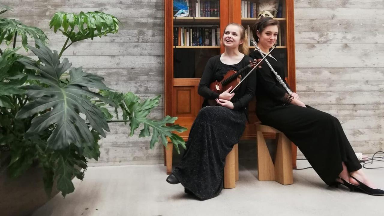 Zwei Frauen in schwarzem Kleid sitzen mit Violine und Querflöte vor einem Bücherschrank. Zwei Frauen in schwarzem Kleid sitzen mit Violine und Querflöte vor einem Bücherschrank.