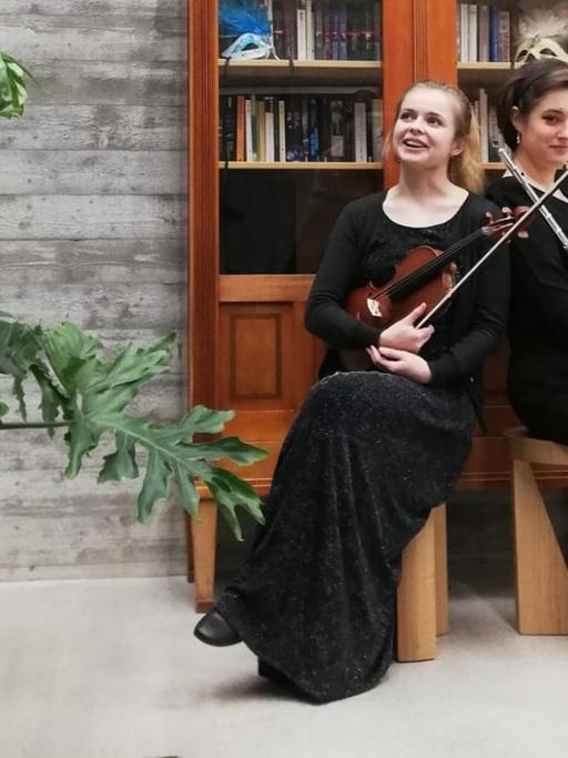 Zwei Frauen in schwarzem Kleid sitzen mit Violine und Querflöte vor einem Bücherschrank.
