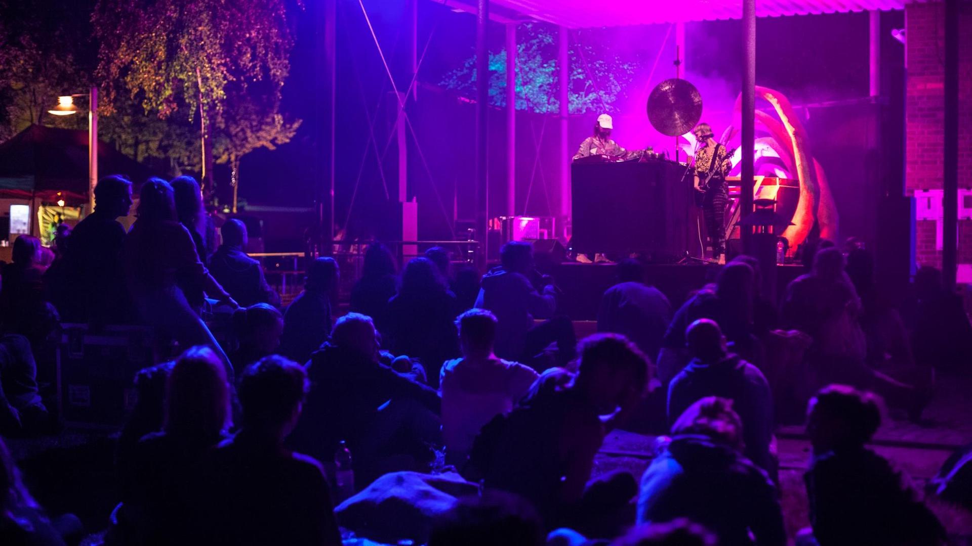 Zuschauer sitzen vor einer in lila und blau angestrahlten Bühne, auf der ein DJ und ein Gitarrist musizieren Zuschauer sitzen vor einer in lila und blau angestrahlten Bühne, auf der ein DJ und ein Gitarrist musizieren