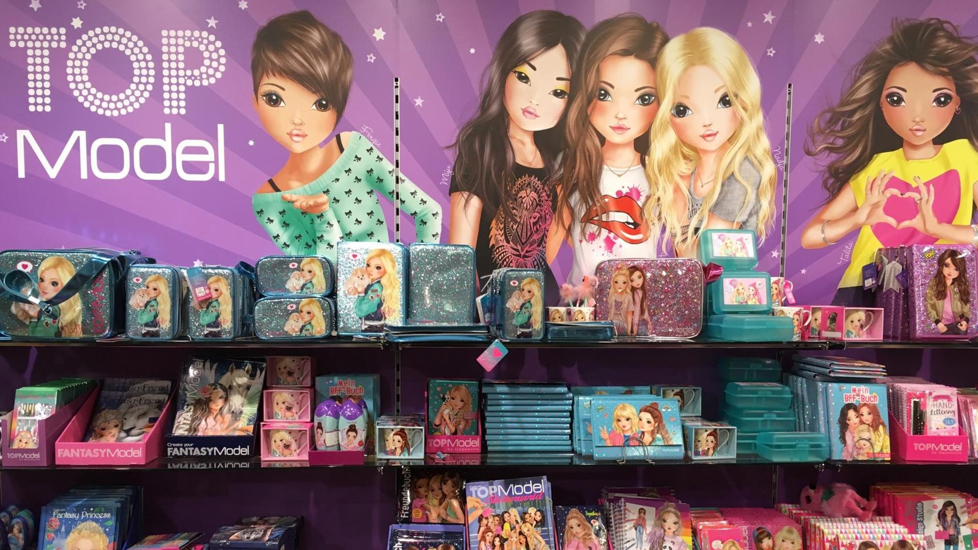 Vier Regalfächer mit pinken und türkisen, auffällig glitzernden Spielwaren. Im Hintergrund hängt ein Plakat mit comichaft gezeichneten Mädchenfiguren und dem Wort "Top Model"