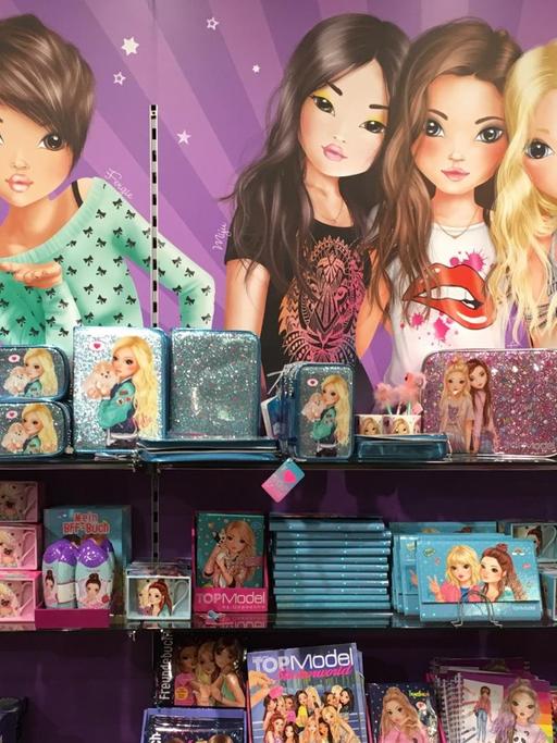 Vier Regalfächer mit pinken und türkisen, auffällig glitzernden Spielwaren. Im Hintergrund hängt ein Plakat mit comichaft gezeichneten Mädchenfiguren und dem Wort "Top Model"