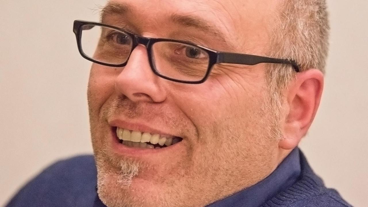 Lutz Neumeier trägt eine schwarze Brille und ein blaues Hemd und lacht an der Kamera vorbei. Lutz Neumeier trägt eine schwarze Brille und ein blaues Hemd und lacht an der Kamera vorbei.