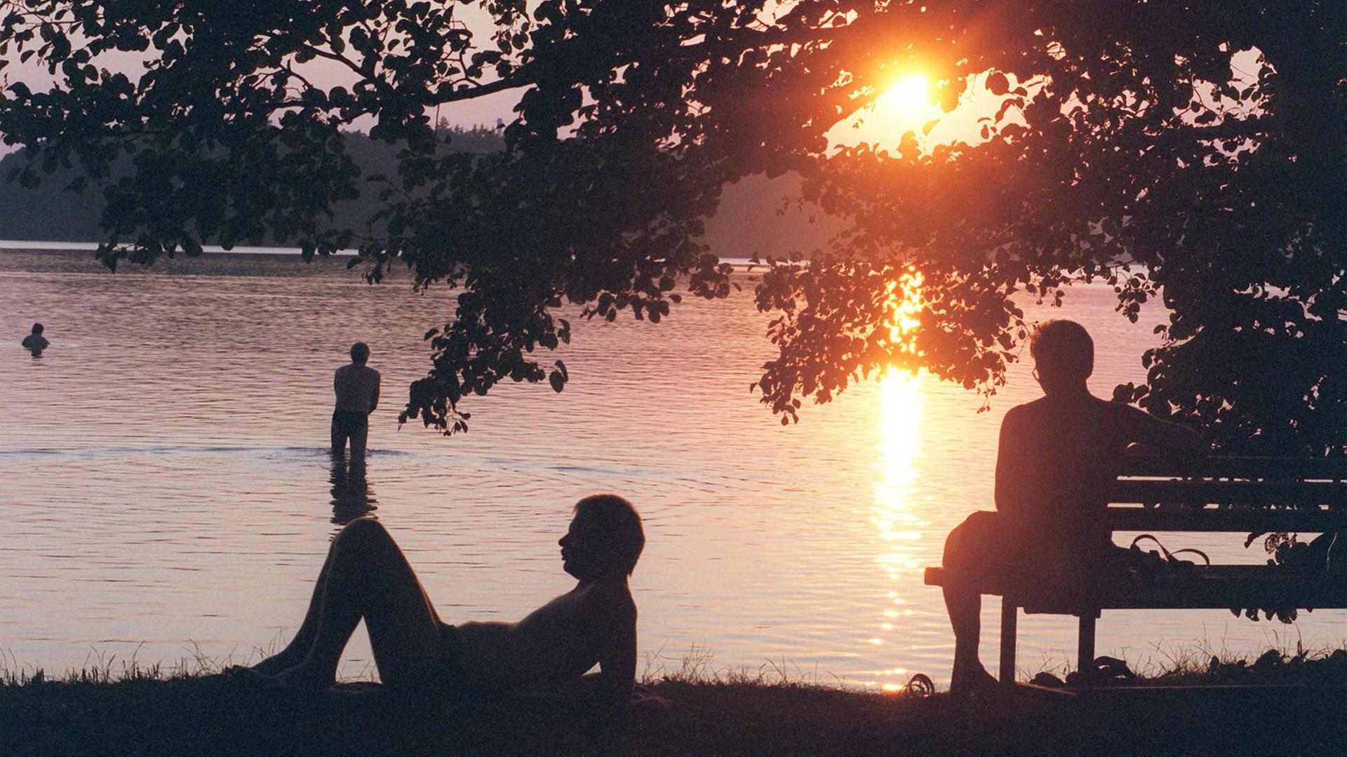 Am Stechlinsee geht die Sonne unter, ein Mensch sitzt am Ufer auf der Bank, einer liegt auf dem Rasen, einer steht im Wasser Am Stechlinsee geht die Sonne unter, ein Mensch sitzt am Ufer auf der Bank, einer liegt auf dem Rasen, einer steht im Wasser