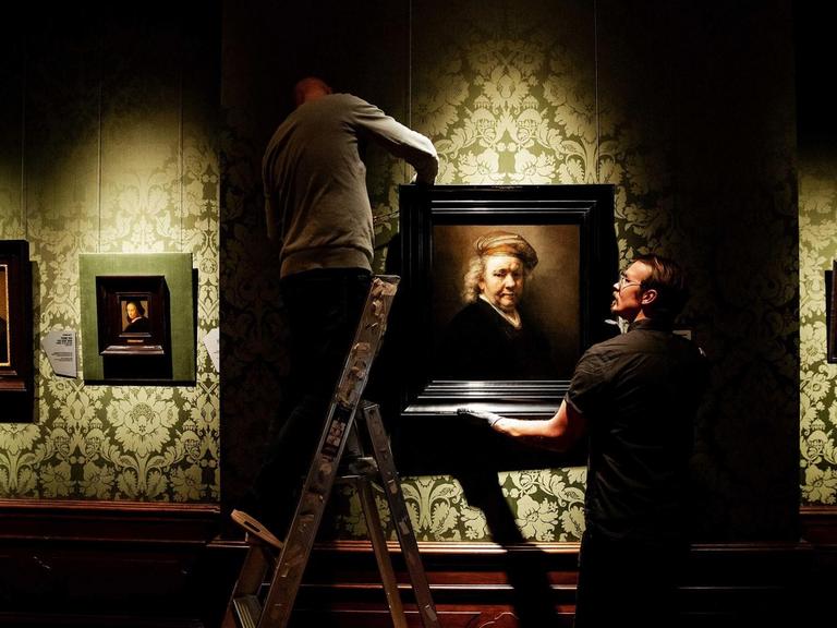 In einer Galerie wird von zwei Mitarbeitern ein Gemälde aufgehängt.