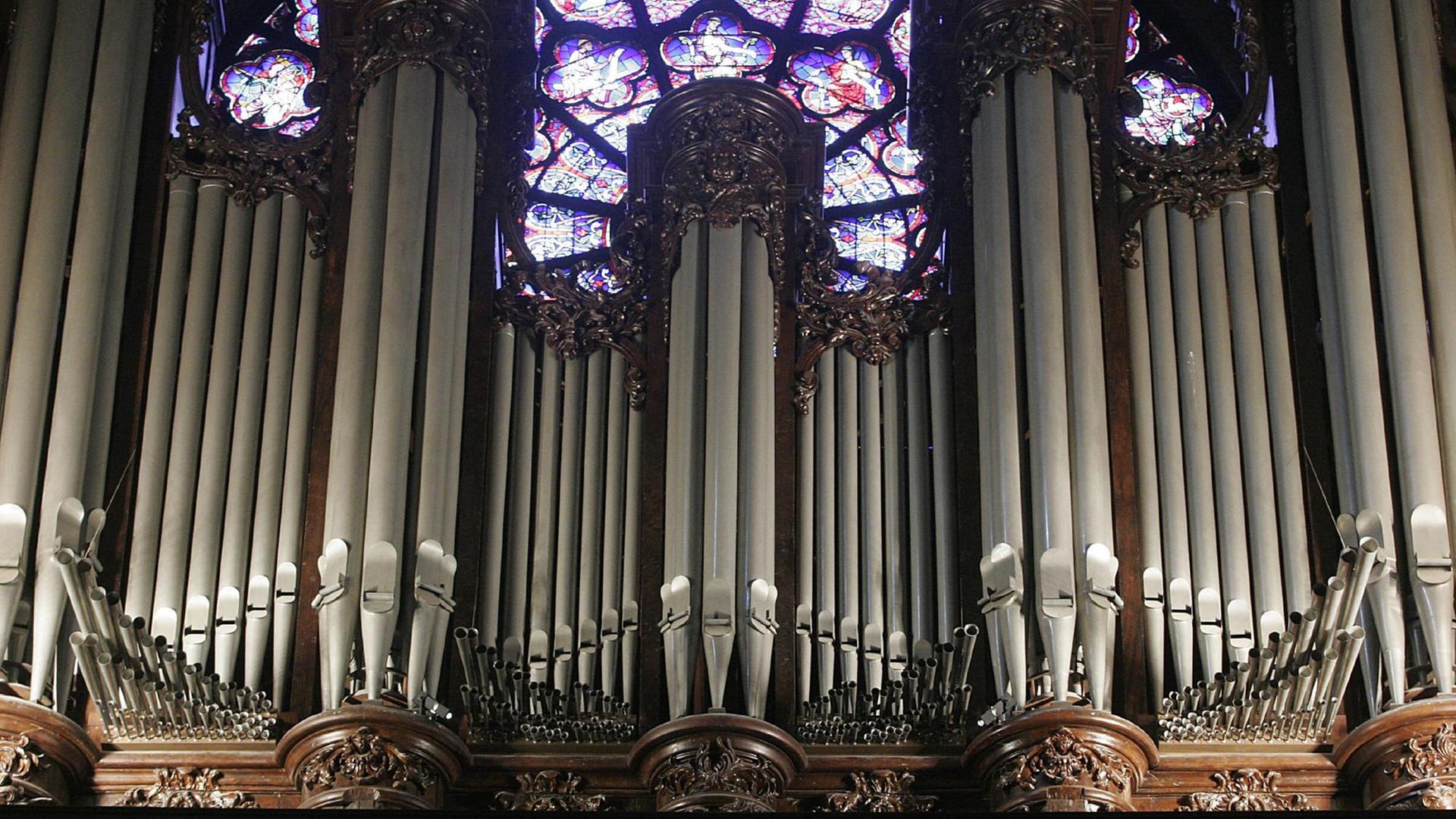 Vor einem rosettenförmigen Kirchenfenster aus blauem, rotem und lilafarbenem Glas stehen viele metallene Orgelpfeifen, eingebundem von reich verziertem Holz. Vor einem rosettenförmigen Kirchenfenster aus blauem, rotem und lilafarbenem Glas stehen viele metallene Orgelpfeifen, eingebundem von reich verziertem Holz.