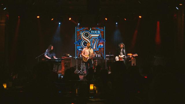 Drei Männer stehen auf einer dunklen Bühne: links der Keyboarder, in der Mitte ein bärtiger Sänger und Gitarrist, rechts der Bassist.
