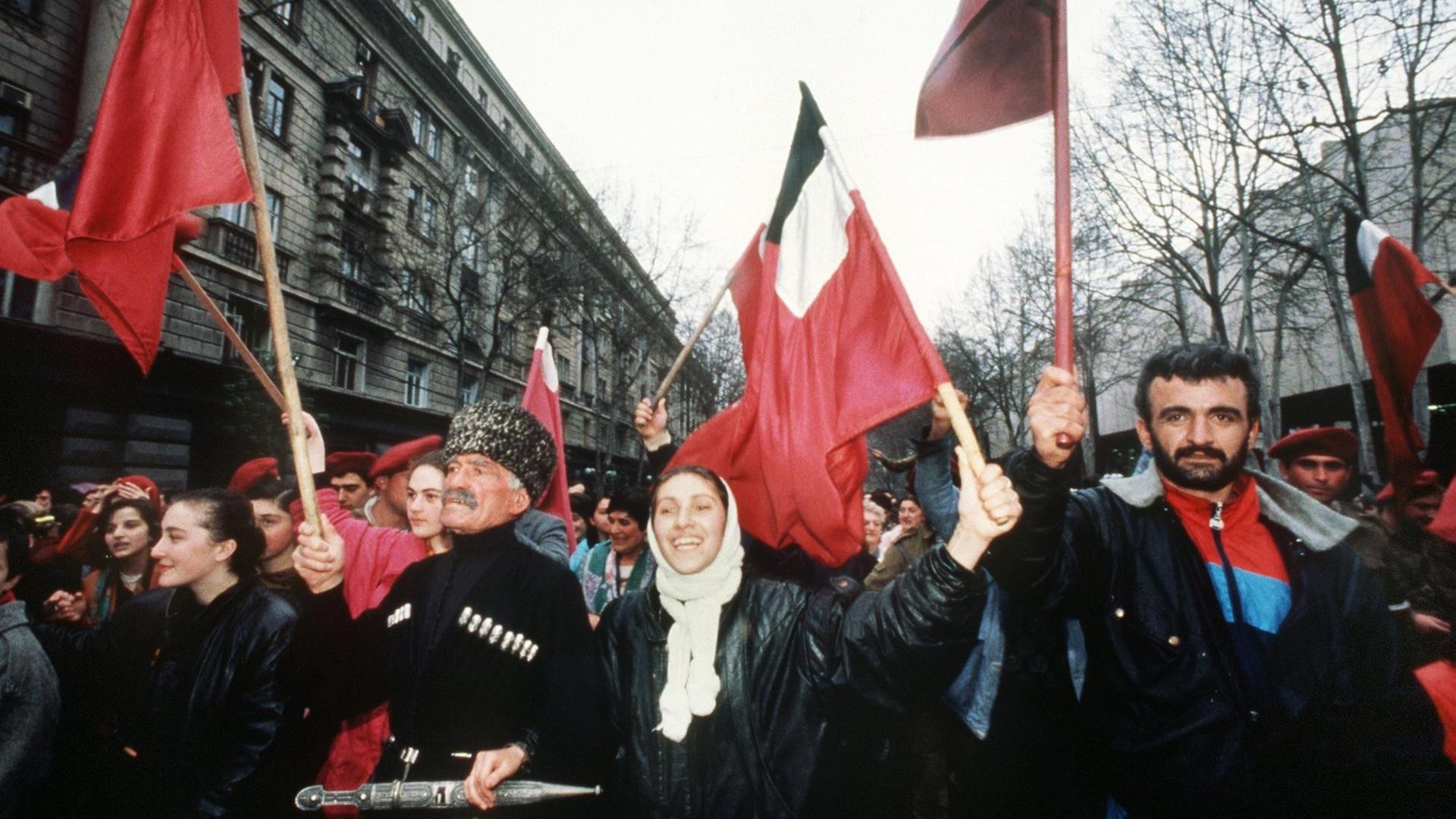 Ein Farbfoto zeigt eine Demonstration Fahnen schwenkender Menschen. in einem historischen Straßenzug. In erster Reihe ein Mann mit traditioneller Fellmütze neben einer gelöst lachenden Frau und einem bärtigen Mann Ein Farbfoto zeigt eine Demonstration Fahnen schwenkender Menschen. in einem historischen Straßenzug. In erster Reihe ein Mann mit traditioneller Fellmütze neben einer gelöst lachenden Frau und einem bärtigen Mann