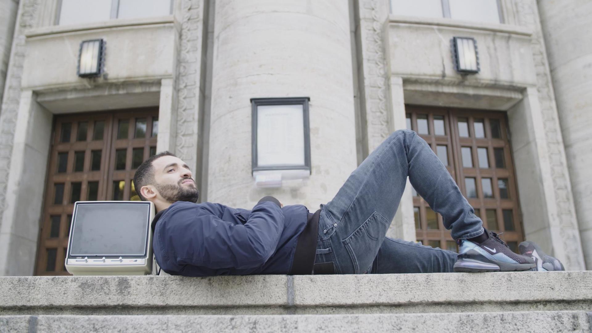 "Kino Kanak. Warum der deutsche Film Migranten braucht": Der Schauspieler Hassan Akkouch liegt vor einem Gebäude auf einer Treppe. Seinen Kopf hat er auf einem alten Fernseher abgelegt und blickt seitlich in die Ferne. "Kino Kanak. Warum der deutsche Film Migranten braucht": Der Schauspieler Hassan Akkouch liegt vor einem Gebäude auf einer Treppe. Seinen Kopf hat er auf einem alten Fernseher abgelegt und blickt seitlich in die Ferne.