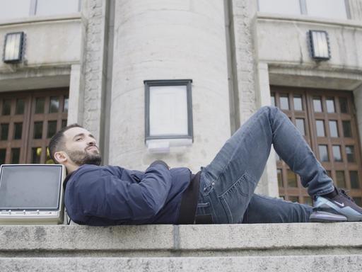 "Kino Kanak. Warum der deutsche Film Migranten braucht": Der Schauspieler Hassan Akkouch liegt vor einem Gebäude auf einer Treppe. Seinen Kopf hat er auf einem alten Fernseher abgelegt und blickt seitlich in die Ferne.
