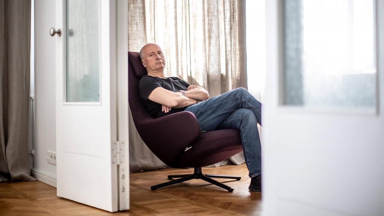 Ein Mann sitzt mit verschränkten Armen auf einem bequemen, großen, modernen Sessel, der durch eine Tür hindurch fotografiert ist.