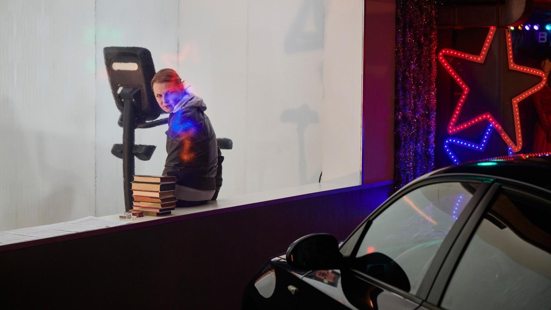 Eine Schauspielerin sitzt auf einen Fenstersims vor einem geparkten Auto. neben ihr liegt ein Bücherstapel. An der Hauswand leuchtet ein großer roter Stern. Eine Schauspielerin sitzt auf einen Fenstersims vor einem geparkten Auto. neben ihr liegt ein Bücherstapel. An der Hauswand leuchtet ein großer roter Stern.