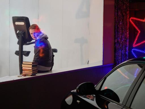 Eine Schauspielerin sitzt auf einen Fenstersims vor einem geparkten Auto. neben ihr liegt ein Bücherstapel. An der Hauswand leuchtet ein großer roter Stern.
